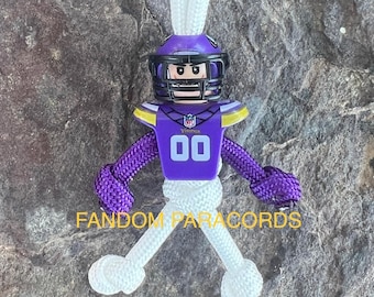 May include: Purple and white paracord keychain with a Minnesota Vikings Lego minifigure. The minifigure is wearing a purple helmet, jersey, and pants with the number 00 on the jersey. The keychain is tied in a knot at the bottom and has a loop at the top.
