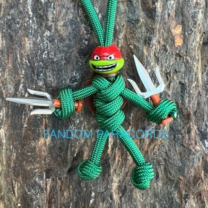 Könnte beinhalten: Eine grüne Paracord-Figur einer Teenage Mutant Ninja Turtle mit einem roten Bandana und einem silbernen Sai. Die Figur hält das Sai in ihrer rechten Hand und hat einen grün-roten Kopf. Die Figur steht auf einer braunen Holzoberfläche. Der Text "RANDOM PARACORDS" ist unten im Bild sichtbar.