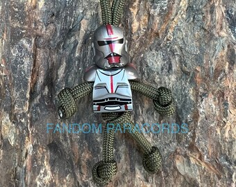 May include: A green paracord keychain with a silver and red Lego-style minifigure of a Star Wars clone trooper. The minifigure has a red stripe down the center of its chest and helmet.