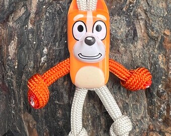 May include: An orange and white cartoon character keychain with a white rope loop. The character has orange arms and a white rope for legs. The keychain is set against a textured, dark brown background.