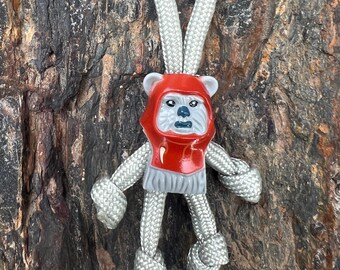 May include: A gray and red Ewok-themed keychain. The figure has a red hood and gray body, arms, and legs made of knotted rope. The keychain loop is made of the same gray rope. The background is a textured brown surface.