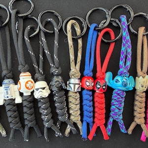 May include: A selection of character keyrings featuring popular characters. The keyrings include Star Wars and Marvel characters, and a Hello Kitty design. Each keyring has a black ring and a braided cord in various colours.