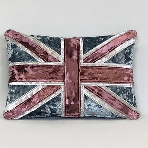 Puede incluir: Una almohada decorativa con un diseño de Union Jack azul y rosa. La almohada está hecha de una tela suave y aterciopelada.