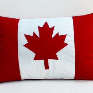May include: A red, white and red cushion with a large red maple leaf in the centre. The cushion is a decorative item with a Canadian flag design.