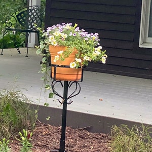 May include: A black metal plant stand with a heart-shaped design supports a terracotta pot filled with white and purple flowers. The stand is on a wooden deck with brown mulch surrounding it.