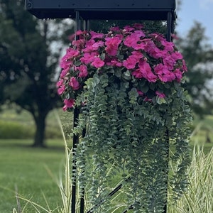 May include: A black mailbox with a red flag is mounted on a black metal stand. A hanging planter with pink flowers and green foliage is attached to the stand.