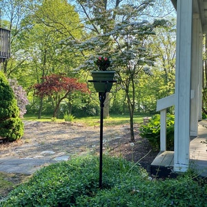 May include: A black metal plant stand with a round planter holding pink and white flowers. The stand is in a garden setting with green grass and trees in the background.