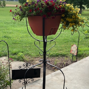 May include: A black metal plant stand with a decorative design holds a terracotta pot filled with a variety of colorful flowers, including red, yellow, and purple.
