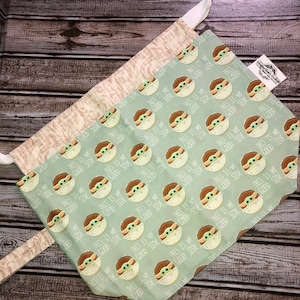 May include: A light green and tan pet bandana with a repeating pattern of a cartoon character and the text "THIS IS MY GOOD SIDE". The bandana is tied with a light tan fabric.