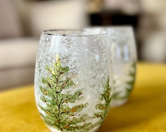 May include: Two clear, stemless wine glasses with a textured, frosted appearance. Each glass features a hand-painted green Christmas tree design with gold accents. The glasses are set on a yellow surface, with a blurred background.