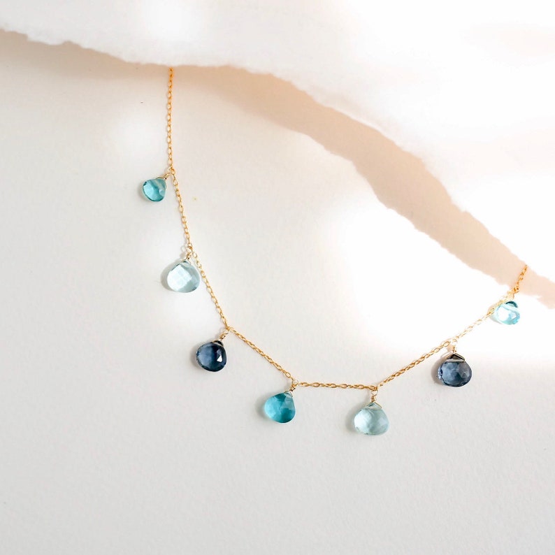 K&ouml;nnte beinhalten: Eine goldene Kettenkette mit tropfenf&ouml;rmigen blauen Edelsteinen, die daran h&auml;ngen. Die Kette liegt auf einem wei&szlig;en Hintergrund.