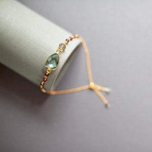 May include: A delicate gold chain bracelet with a green gemstone pendant and small red beads. The bracelet has a sliding clasp for adjustable sizing.