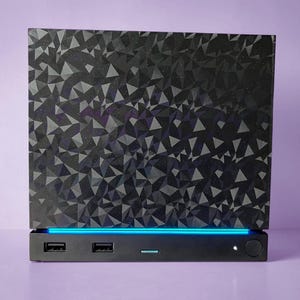 May include: A black rectangular electronic device featuring a top panel with a geometric, faceted, dark gray pattern. The base includes two USB ports, a thin horizontal blue light strip, a small indicator light, and a circular power button.