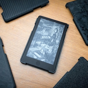 Könnte beinhalten: Ein schwarzer E-Reader mit einem Bildschirm, der eine Graustufen-Illustration einer Straßenszene zeigt, umgeben von vier verschiedenen schwarzen Schutzhüllen mit unterschiedlichen Texturen wie Carbonfaser, Camouflage und matten Oberflächen.