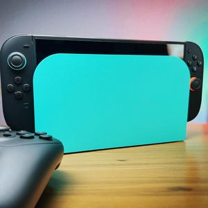 Peut inclure: Une console de jeu noire avec un support turquoise est présentée sur une surface en bois. Une manette noire est au premier plan. La console a des contrôleurs noirs avec des boutons et un écran. Une petite plante est en arrière-plan. Idéal pour les jeux.