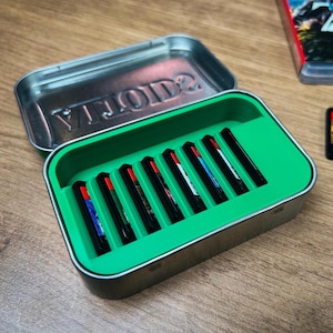 May include: An open metal tin with a green interior containing several black game cartridges with red detailing. A game cartridge and case are also visible. The cartridges are for the Nintendo Switch console.