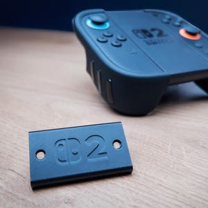 May include: A black Switch controller with blue and orange buttons, and a separate black plate with the text "D2". The controller is designed for gaming and has a sleek, modern design. The controller is on a wooden surface.