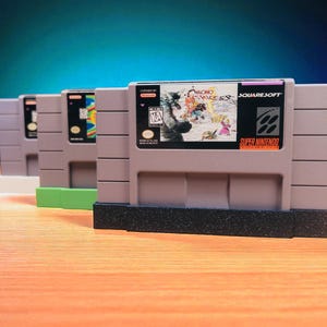 May include: Four gray Super Nintendo game cartridges are displayed, with the front-most cartridge featuring the game "Chrono Trigger" by SquareSoft. The cartridges are arranged in a row, with the first three resting on a black base and a green base.