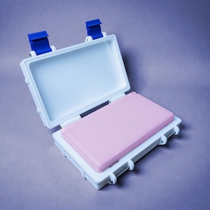 May include: An open, white, rectangular case with a pink interior and blue latches. The case is on a purple surface. The top half of the case is open, revealing the pink interior. The case is designed to protect items.
