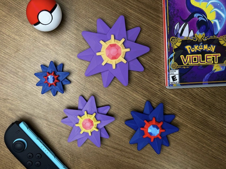 A collection of Pokemon-themed items. There is a red and white Poke Ball, a Nintendo Switch controller, and a Pokemon Violet game case. Also, there are five star-shaped decorations in purple and blue with yellow and red center designs. The items are on a wooden surface.