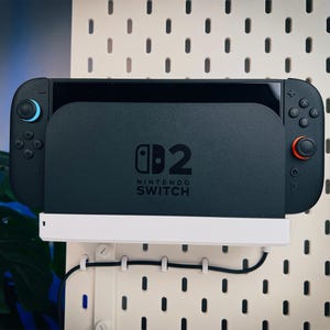 以下が含まれることがあります： 白いペグボードに取り付けられた、黒いNintendo Switchコンソール。コンソールにはNintendo Switchのロゴと白いベースがあります。左側のコントローラーには青いジョイスティック、右側にはオレンジ色のジョイスティックが付いています。