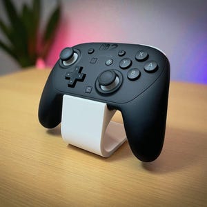 May include: A black Nintendo Switch Pro controller with white accents, resting on a white stand. The controller features a directional pad, analog sticks, and various buttons. The stand is curved and designed to hold the controller upright.