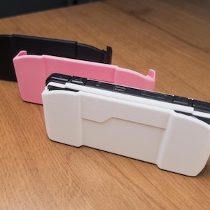 以下が含まれることがあります： 携帯ゲーム機用の保護ケース3つ。ケースは黒、ピンク、白です。白いケースが手前にあり、黒い上部と幾何学模様のデザインです。ケースは木製の表面にあります。