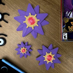 Three purple Staryu Pokemon figures with gold and red accents are displayed on a wooden surface. The figures vary in size, with one large and two smaller ones. Also present are black Unown figures and a Nintendo Switch console. A Pokemon Violet game case is visible.