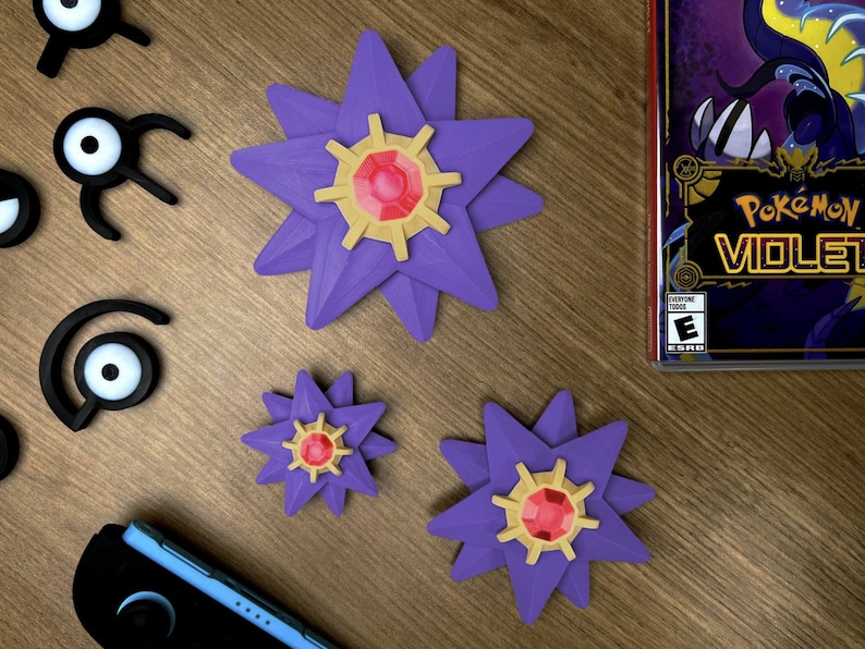 Three purple Staryu Pokemon figures with gold and red accents are displayed on a wooden surface. The figures vary in size, with one large and two smaller ones. Also present are black Unown figures and a Nintendo Switch console. A Pokemon Violet game case is visible.