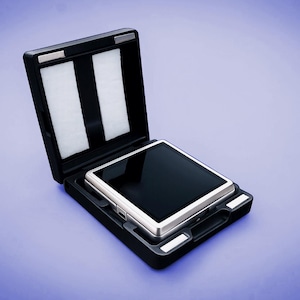 May include: A silver and black electronic device rests inside an open black case with white padding. The case has a handle and is set against a gradient blue background. The device has a reflective black screen.
