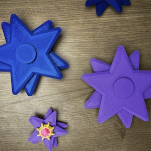 Several star-shaped fidget spinners in various colors are displayed on a wooden surface. There are two blue spinners, one purple spinner, and one purple spinner with a yellow and pink center. Each spinner has a circular center and multiple points.