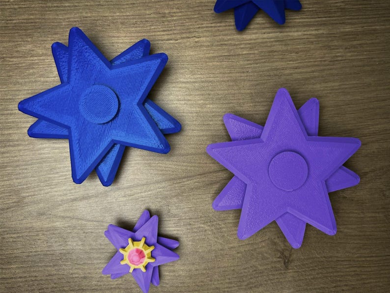 Several star-shaped fidget spinners in various colors are displayed on a wooden surface. There are two blue spinners, one purple spinner, and one purple spinner with a yellow and pink center. Each spinner has a circular center and multiple points.