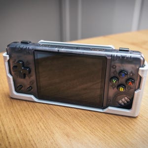 Puede incluir: Consola de juegos portátil en una carcasa de plástico transparente, sujeta en un soporte blanco impreso en 3D. La consola tiene una pantalla negra, un mando direccional y botones de colores. El dispositivo está sobre una superficie de madera.