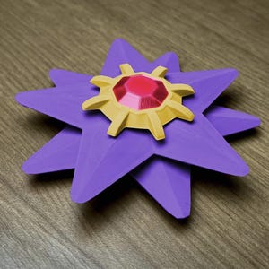 A purple, star-shaped decorative item with a gold and red center. The star has eight points, each with a folded appearance. The center features a gold, sun-like design with a large, red, gem-like stone in the middle. The item is resting on a wooden surface.