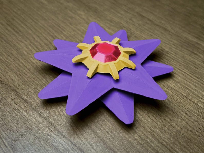 A purple, star-shaped decorative item with a gold and red center. The star has eight points, each with a folded appearance. The center features a gold, sun-like design with a large, red, gem-like stone in the middle. The item is resting on a wooden surface.