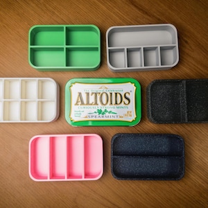 May include: A collection of small, compartmented trays in green, grey, white, pink, and black, next to a green Altoids Spearmint tin. The trays are designed for organizing small items, with multiple sections.