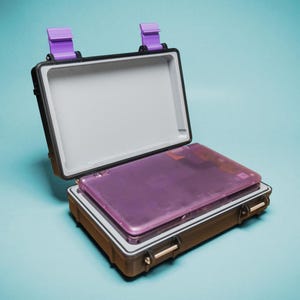 May include: A brown waterproof case, open to reveal a purple laptop. The case has black trim, purple latches, and gold-coloured clasps. The laptop is a translucent purple colour. The background is a solid light blue.