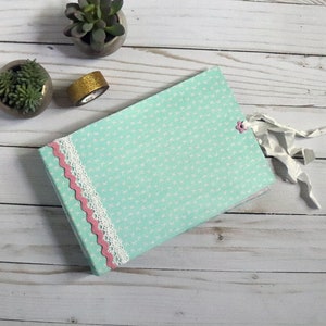 May include: A light blue journal with a white cross pattern, accented with pink and white lace trim. A small pink flower and white ribbon are attached. Two small succulent plants in gray pots and a roll of gold glitter tape are also visible.