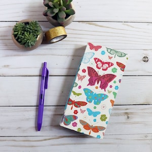 May include: A white journal with a butterfly pattern, a purple pen, two small potted succulents, and a roll of gold glitter tape are arranged on a white wooden surface. The journal features colorful butterflies and flowers.