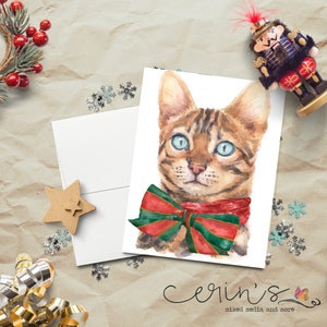 May include: A watercolor painting of a brown tabby cat wearing a red and green bow tie. The cat has blue eyes and is looking at the viewer. The background is white.