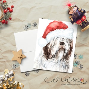 May include: A white greeting card featuring a watercolor illustration of a shaggy dog wearing a red and white Santa hat. The card is positioned on a textured surface surrounded by holiday decorations including a wreath, a nutcracker, and ribbons.