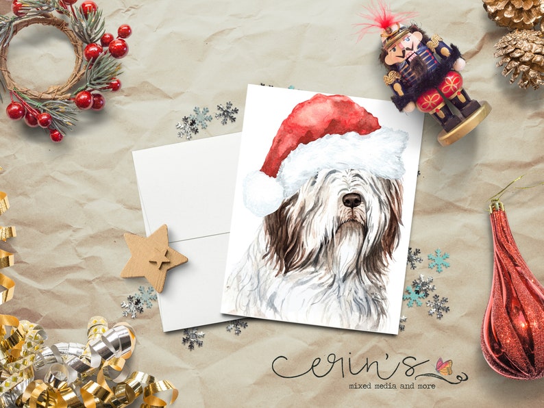 May include: A white greeting card featuring a watercolor illustration of a shaggy dog wearing a red and white Santa hat. The card is positioned on a textured surface surrounded by holiday decorations including a wreath, a nutcracker, and ribbons.