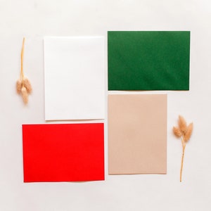 May include: Four blank cards in different colors: red, white, green, and beige. The cards are arranged in a grid pattern on a white background. Two sprigs of dried pampas grass are placed on either side of the cards.