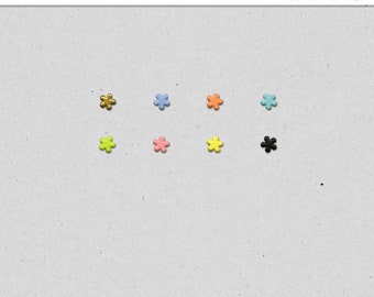 May include: Assortment of tiny flower-shaped brads in various colors, including gold, blue, orange, green, pink, yellow, and black. The brads are arranged in two rows of four. The text "Tiny Brads Vol. 1: Flowers" is at the top of the image.