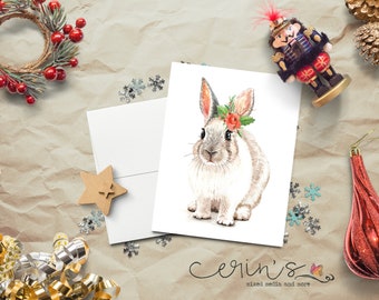 May include: A white greeting card featuring a watercolor illustration of a light brown and white rabbit wearing a festive holly and red berry wreath on its head. The card is positioned against a textured background with holiday decorations.
