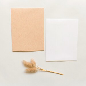 May include: A rectangular light brown kraft paper card and a rectangular white card positioned side by side, accompanied by a small cluster of three dried, fluffy, light tan bunny tail grass stems with thin, light brown stalks.