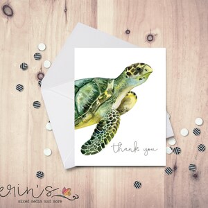May include: A watercolor greeting card featuring a green sea turtle with yellow accents. The card is inside a white envelope, with the words "thank you" in script. Scattered confetti and a wooden background.