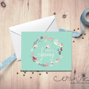 May include: A light blue card with a floral wreath and two birds. The wreath is made of pink and white flowers, green leaves, and brown branches. The text "Happy Spring" is written in white cursive lettering inside the wreath. The card is on a wooden surface.
