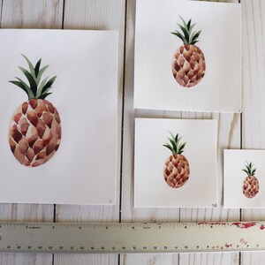 May include: Four different sizes of watercolor pineapple prints on white paper. The pineapple is brown with a green top. The prints are labeled with the sizes 16 and 28.