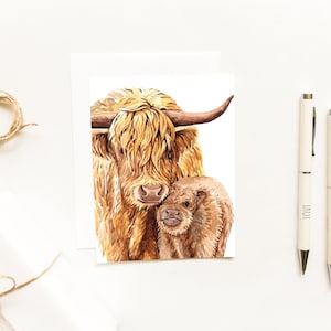 May include: A greeting card featuring a watercolor illustration of a shaggy Highland cow and its calf. The card is accompanied by two white pens with the words LOVE and HOPE printed on them, a white envelope, and a small coil of twine.